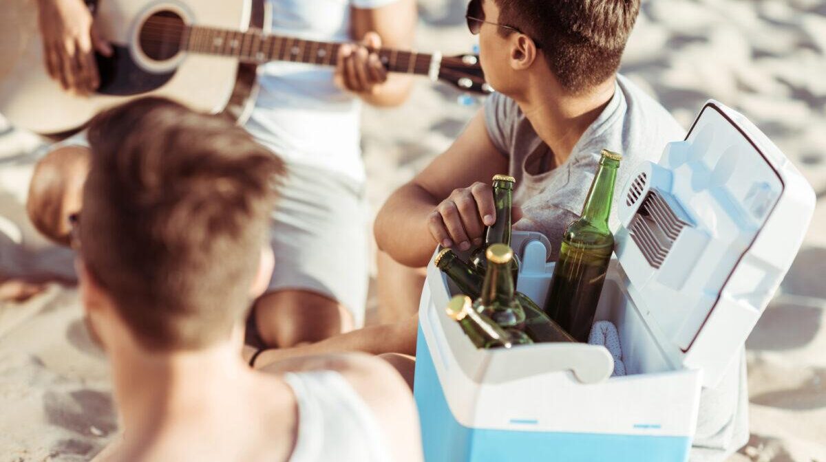 Junge Leute sitzen mit einer der besten Kühlboxen am Strand, trinken ein Bier und machen zusammen Musik.